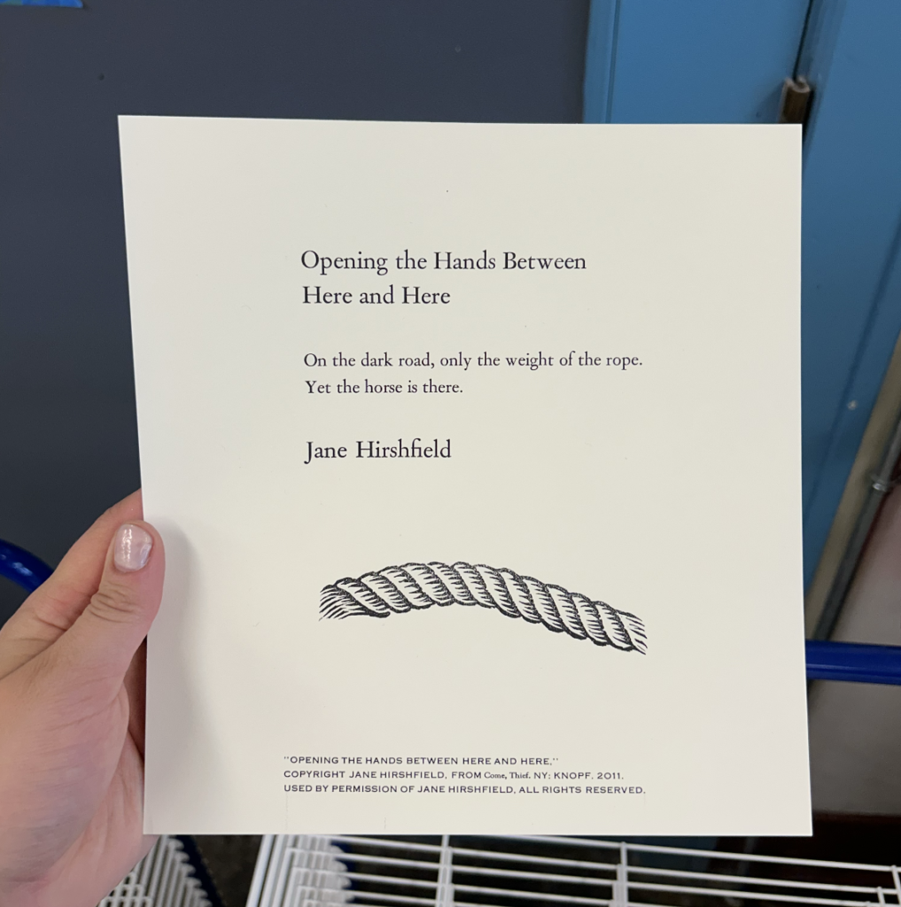 Chloë's hand holds up a print of "Opening the Hands Between Here and Here" by Jane Hirshfeld with an image of a rope at the bottom. 